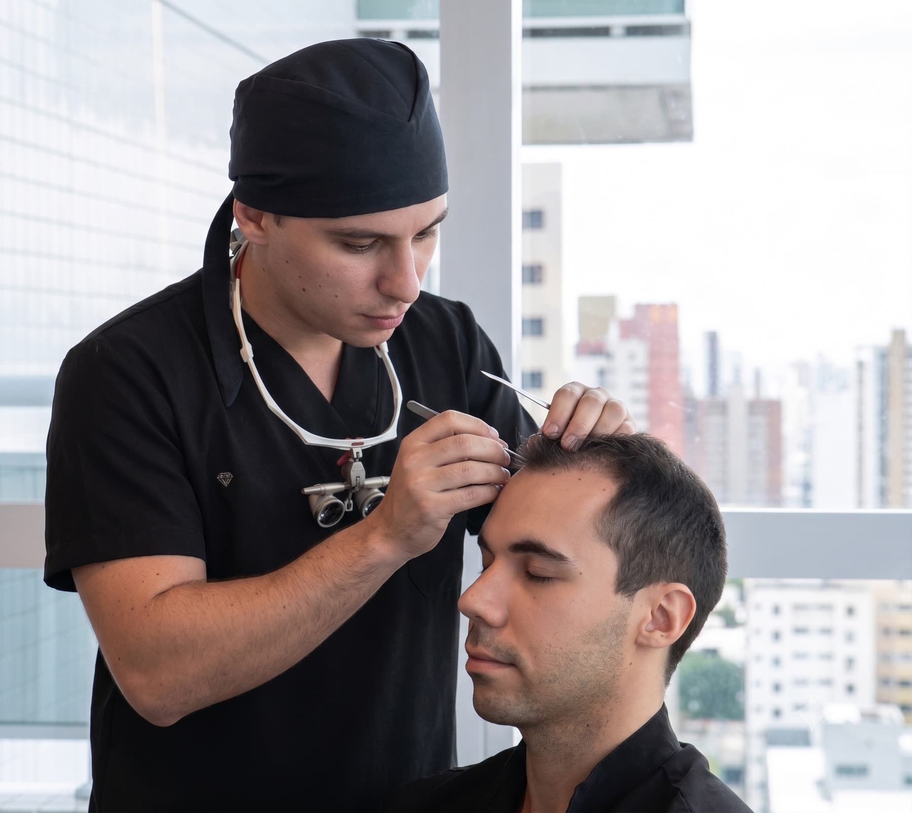 Dr. Lucas Moura em ato cirúrgico: uniforme preto, lupas operatórias, atendendo paciente sentado em ambiente clínico com luz natural e vista urbana.