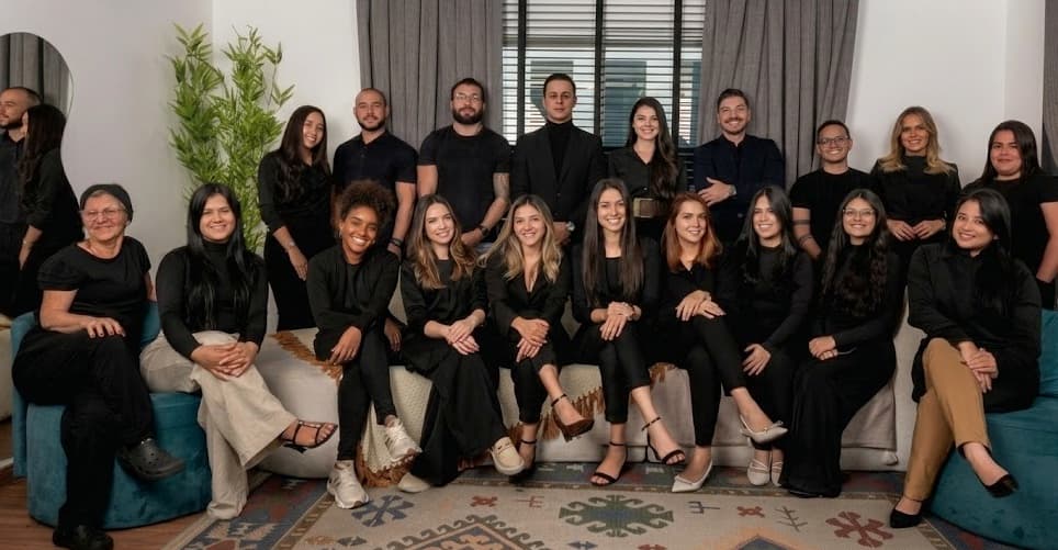 Equipe consolidada Dr. Lucas Moura: vinte profissionais com uniformes pretos reunidos em foto institucional, sentados em sofá e poltrona com tapete kilim de motivos geométricos em ambiente claro com cortinas e planta natural ao fundo.