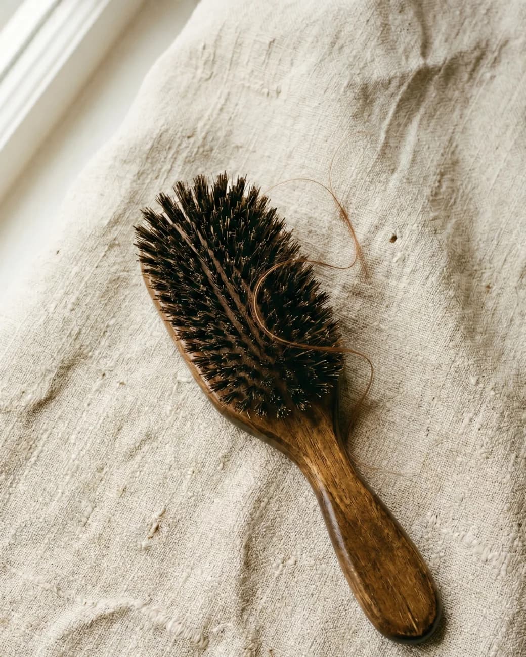 Escova de cabelo de madeira com alguns fios sobre tecido de linho natural à luz suave de janela, em paleta creme.