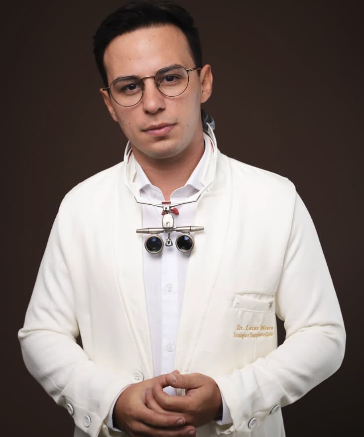 Dr. Lucas Moura em retrato de pé, jaleco branco com lupa binocular ao colo, fundo marrom em iluminação editorial.