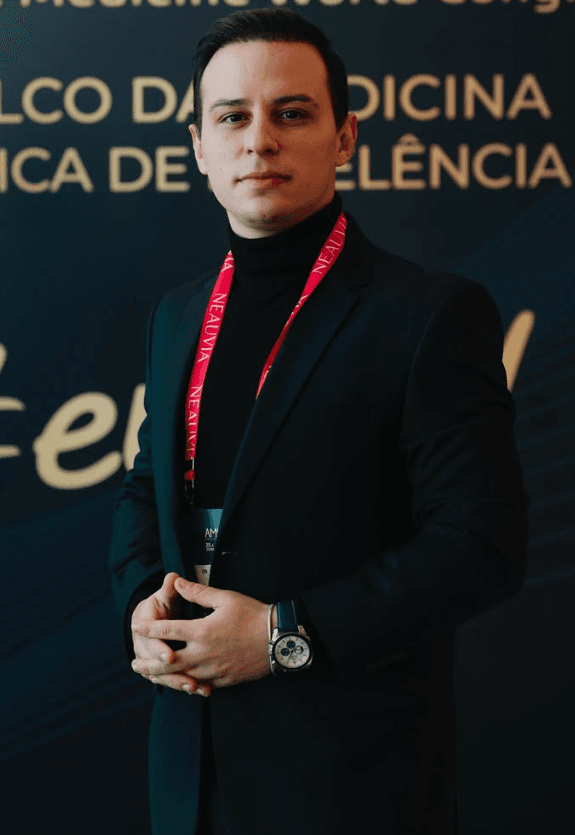 Retrato em pé do Dr. Lucas Moura em terno preto e gola alta, mãos sobrepostas em postura institucional, cordão e credencial de congresso médico internacional ao pescoço, expressão serena em ambiente de evento profissional.